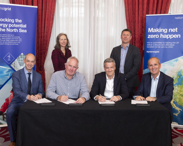 6 people stood behind a table, smiling at the camera and signing a contract