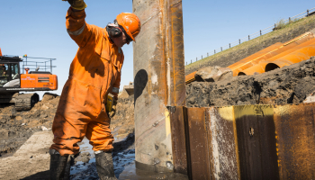 Piles being driven into the ground on a construction site with man in orange PPE stood next to them